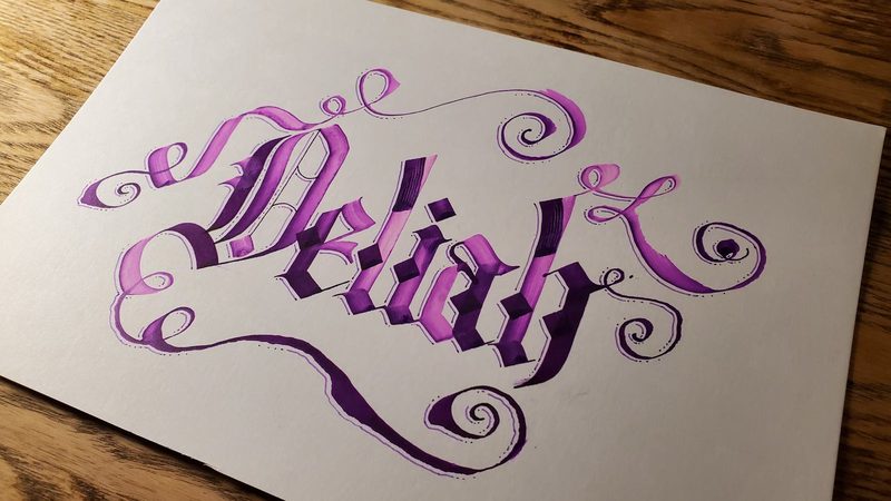 The name “Deliah” hand-lettered in blackletter calligraphy, shaded in violet and deep purple with looping flourishes, on cream paper resting on a wooden table.